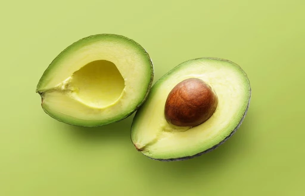 An avocado cut in half, showing the bright green flesh and the large brown pit in the center, set against a solid green background.