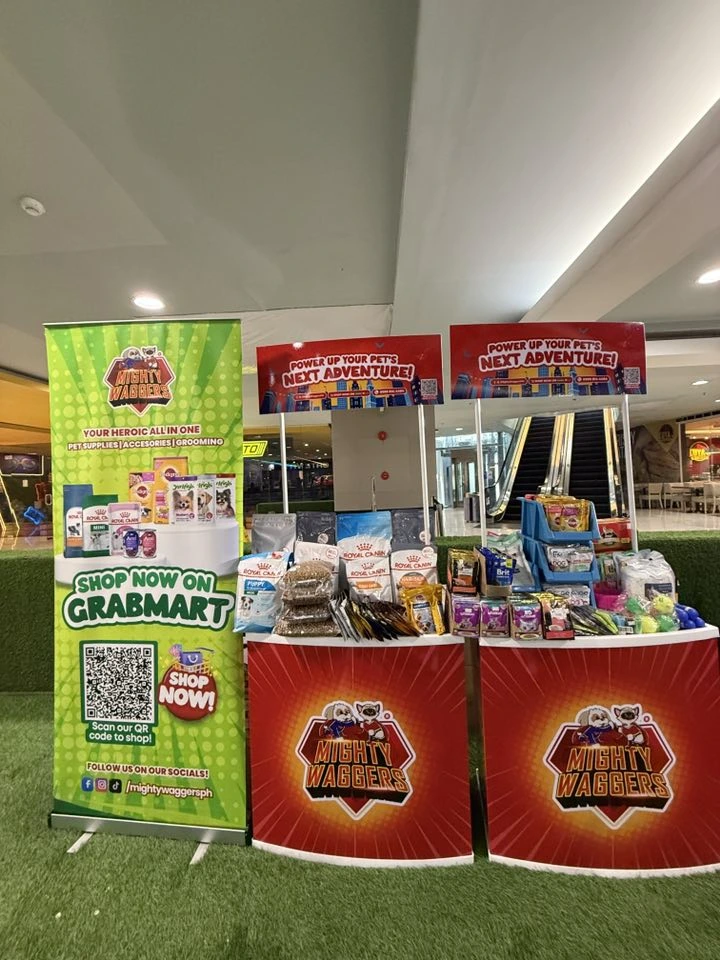 A full view of the Mighty Wagbers vendor booth selling pet supplies, featuring a tall banner with a QR code for shopping on GrabMart and products displayed on two tables.