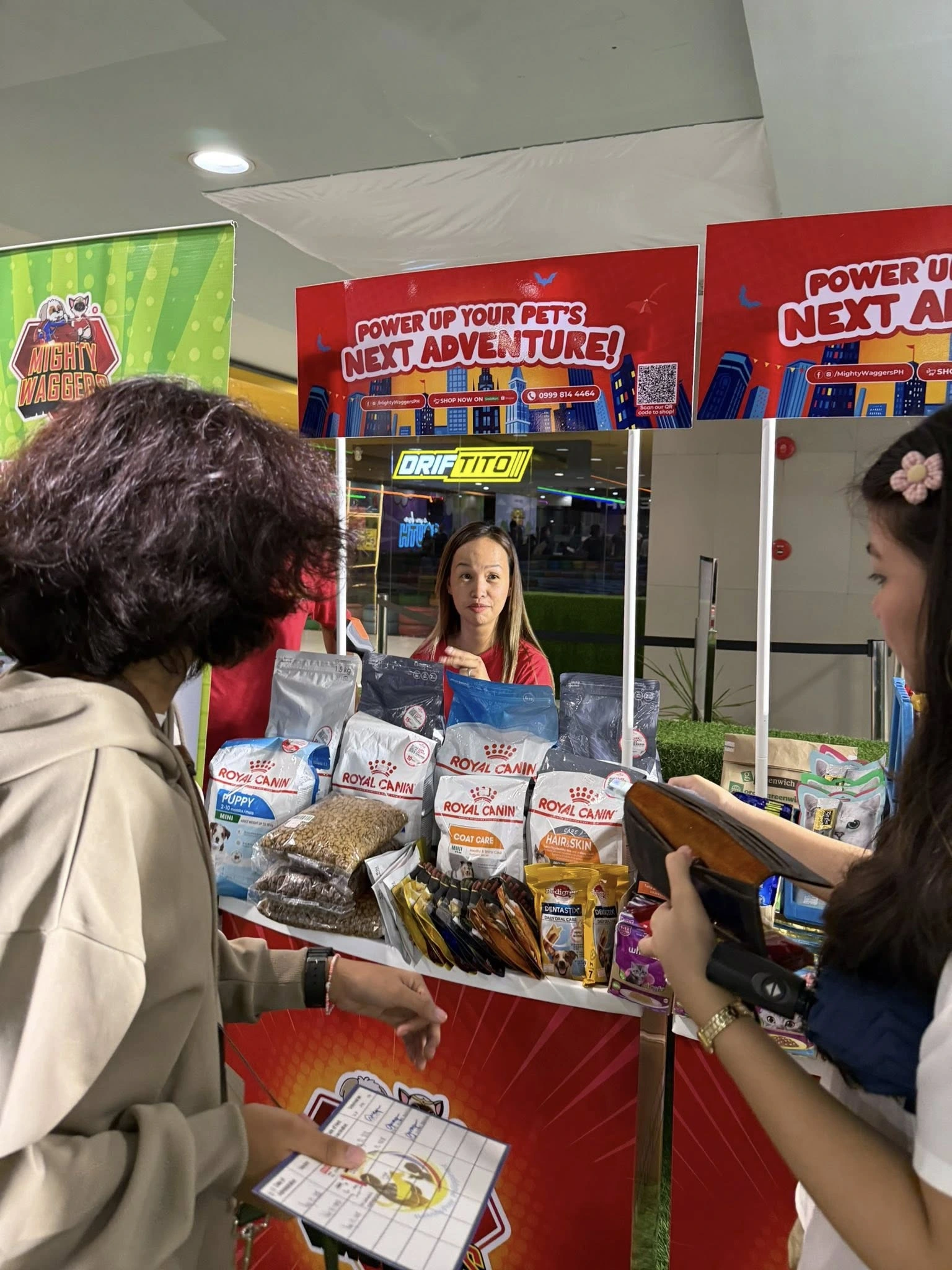 A full view of the Mighty Wagbers vendor booth selling pet supplies, featuring a tall banner with a QR code for shopping on GrabMart and products displayed on two tables.