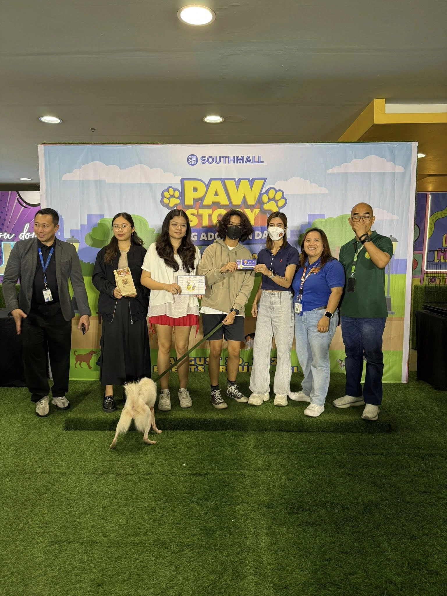 A group of seven people and a small white dog stand on a grassy stage in front of a blue banner that reads "SM Southmall Paw Story Dog Adoption Day.