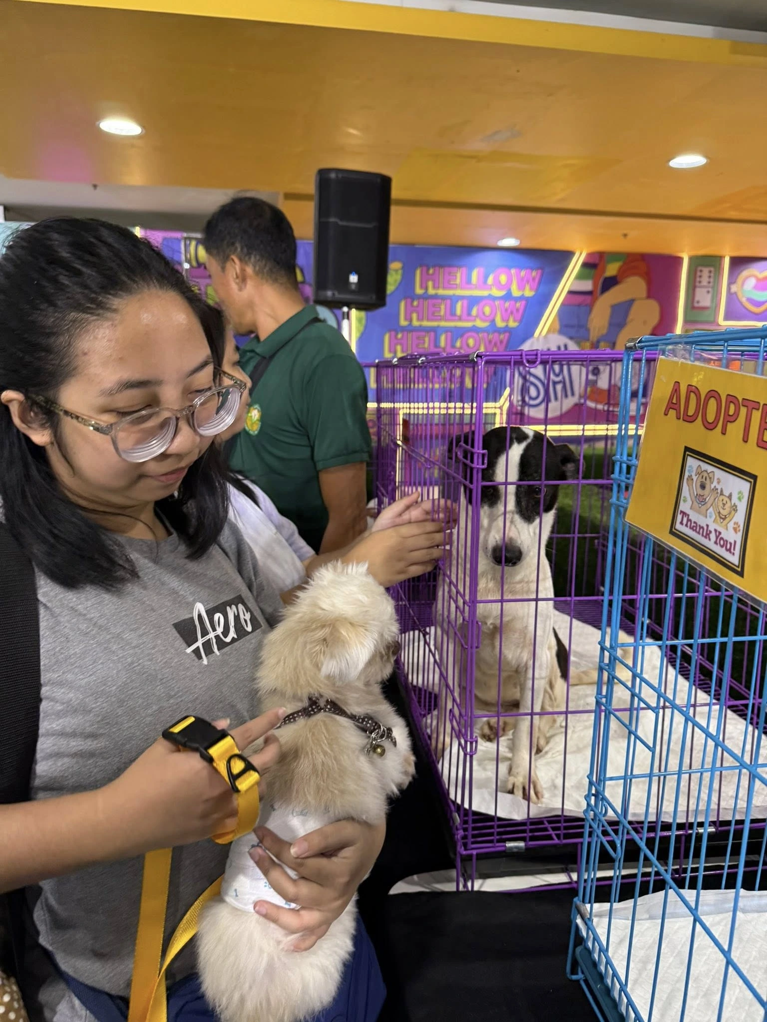 A woman holding a small, light-colored puppy with a yellow leash, while a black-and-white dog sits in a purple adoption cage with an "ADOPTED" sign on it.