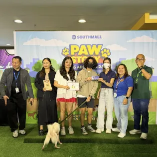 Group of people on a stage at SM Southmall presenting a dog adoption certificate, part of a "PAW STORY Dog Adoption Day" event.
